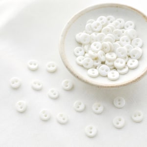 May include: A collection of small, round, white buttons, each featuring two central holes. A large group of these buttons is contained within a shallow, off-white ceramic bowl, while several individual buttons are scattered around the base.