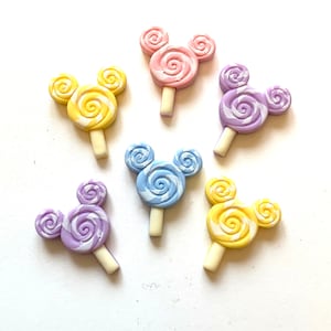 May include: Six colorful, Mickey Mouse-shaped lollipops. The lollipops are in shades of pink, yellow, blue, and purple, with white swirl designs. Each lollipop has a white stick.