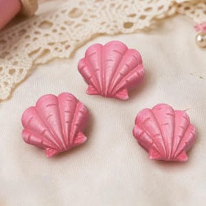 May include: Three small, pink, shell-shaped buttons with a ridged, fan-like texture. Each button features a scalloped edge and a smooth, matte finish, designed with a classic bivalve mollusk appearance for use as decorative garment fasteners.