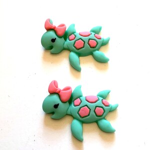 May include: Two sea turtle embellishments. Each turtle is light teal with pink accents on the shell and a pink bow. The turtles have black eyes and are on a white background.