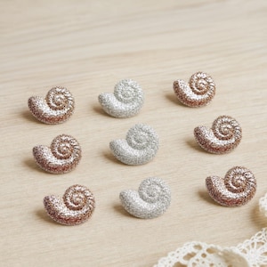 May include: A collection of eight small, snail-shaped buttons arranged in three rows. The buttons feature a textured, glittery finish in alternating rose gold and silver colors, each displaying a distinct spiral shell pattern on the surface.