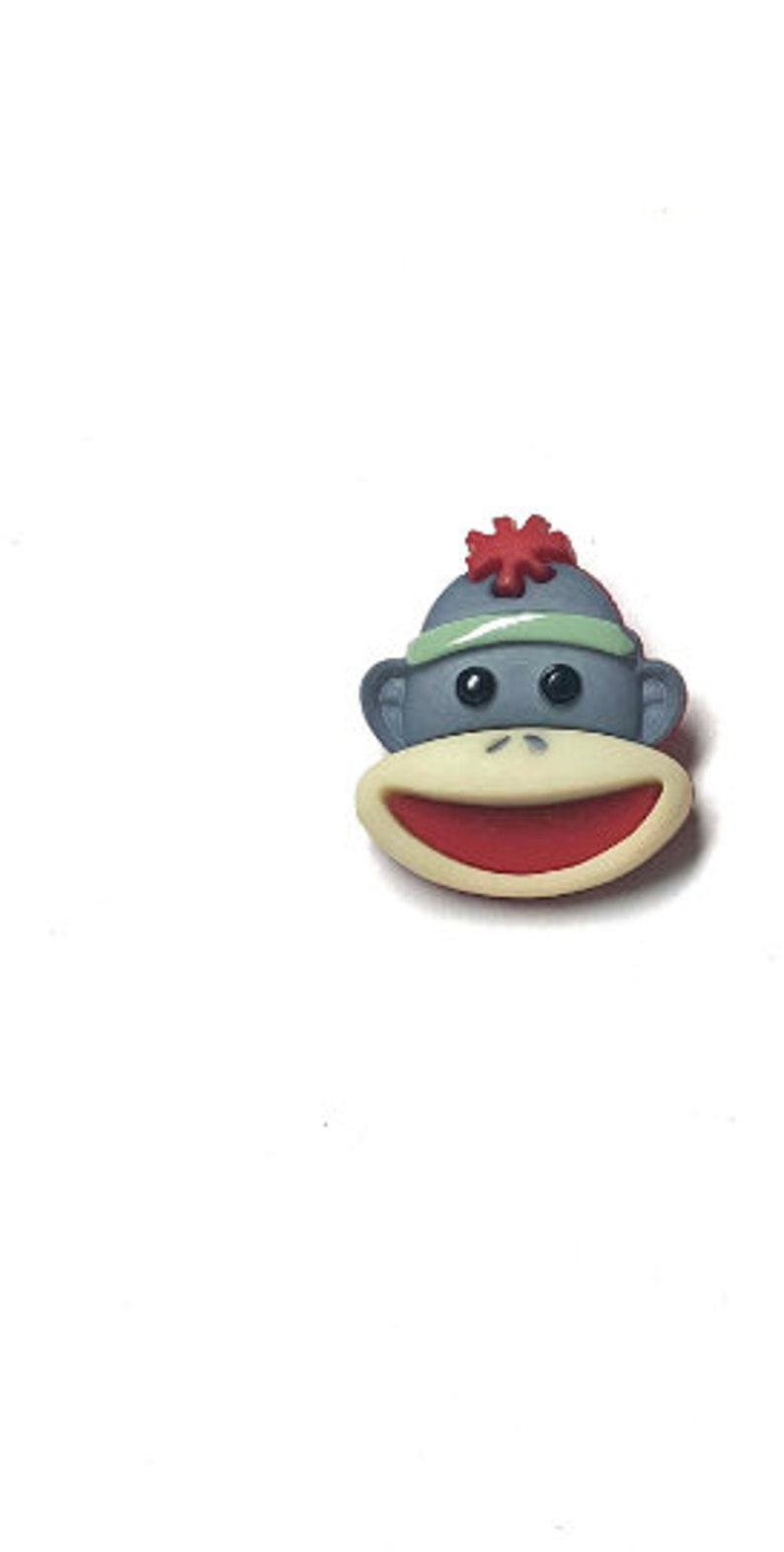 Puede incluir: Una cara de mono de dibujos animados con una boca roja y una cabeza gris. El mono lleva un sombrero gris con un pomp&oacute;n rojo y una diadema verde. La cara del mono es de color beige claro.