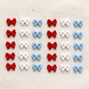 May include: A collection of thirty small bow-shaped buttons arranged in a grid. The buttons are colored in red, white, and light blue. Each button features two holes in the center for sewing, with a smooth, glossy finish on the surface.
