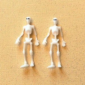 May include: Two small, white plastic skeleton figurines with black dot eyes. The skeletons are standing upright with arms outstretched. The background is a textured, light tan surface.