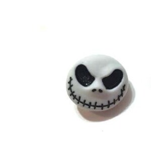 May include: A white, round decorative button featuring the face of Jack Skellington from The Nightmare Before Christmas. The button has black eyes and a stitched smile. The button is on a white background.
