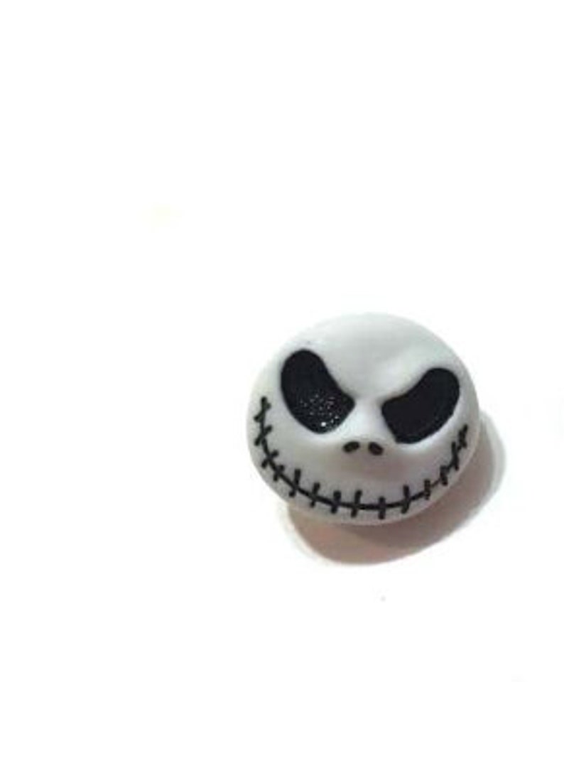 May include: A white, round decorative button featuring the face of Jack Skellington from The Nightmare Before Christmas. The button has black eyes and a stitched smile. The button is on a white background.