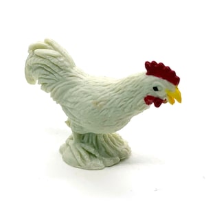 May include: A small, off-white figurine of a rooster. The rooster has a red comb and wattle, a yellow beak, and detailed feathering. The figurine is standing on a textured base, and the overall design is highly detailed.