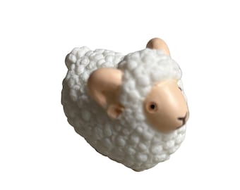 May include: A small, white sheep figurine with a textured, wool-like body. The sheep has a light tan face, ears, and horns, with small, dark eyes. The figurine is a charming decorative item.