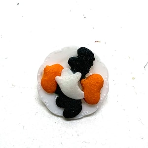 May include: A small, white, scalloped-edge plate holds Halloween-themed sprinkles. The sprinkles include orange pumpkins, black bats, and white ghosts. The plate is set against a white background.