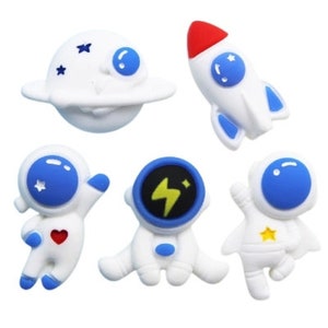 May include: A collection of white and blue space-themed embellishments. Includes a planet with stars, a rocket with a red tip, and three astronaut figures. The astronaut figures have blue visors and various details like a heart and a star.