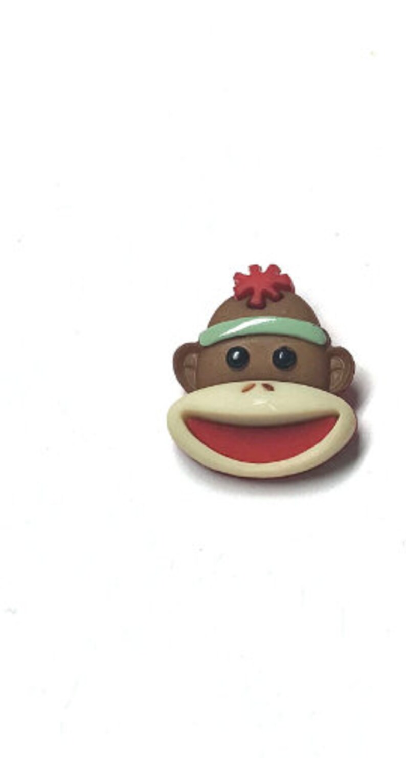 Puede incluir: Primer plano de un bot&oacute;n con la cara de un mono Paul Frank. El bot&oacute;n presenta una cabeza de mono marr&oacute;n con una diadema verde claro y una flor roja. El mono tiene una boca color crema con el interior rojo y ojos negros.