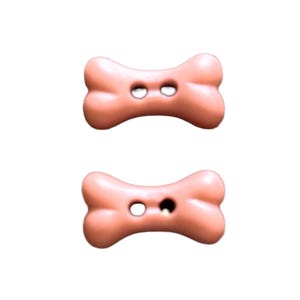 May include: Two light pink buttons shaped like dog bones. Each button features a smooth, rounded surface with two centered holes for attachment. The buttons have a uniform matte finish and a distinct, symmetrical bone-like silhouette.