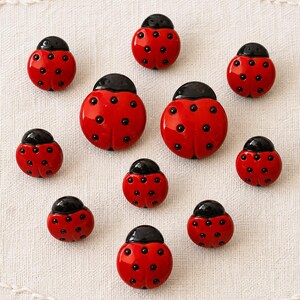 May include: A collection of ten ladybug-shaped buttons, each featuring a glossy red body with black spots and a black head. The buttons vary slightly in size, with two larger ones positioned centrally and eight smaller ones arranged around them.