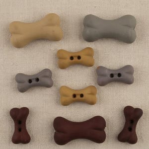 May include: A collection of nine bone-shaped buttons in various shades of brown and gray. Some buttons feature two central holes for sewing, while others are solid. The buttons are arranged in a scattered pattern, displaying smooth surfaces.