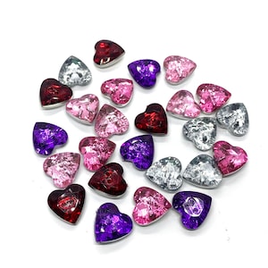 May include: A collection of heart-shaped embellishments in various colors, including red, pink, purple, and silver. The embellishments have a faceted, gem-like appearance and are arranged on a white surface. These decorative hearts are suitable for crafts.