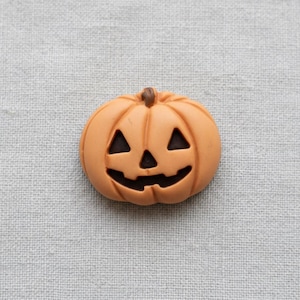 May include: A light orange pumpkin-shaped button featuring a carved jack-o'-lantern face with two triangular eyes, a triangular nose, and a wide, jagged mouth. The surface is smooth with a small brown stem at the top center of the pumpkin.