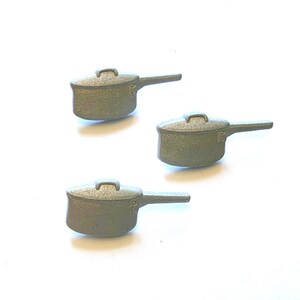 May include: Three small, olive-green saucepans with lids and long handles are arranged on a white background. The saucepans have a textured surface and a simple, classic design, suitable for miniature cooking or decorative purposes.