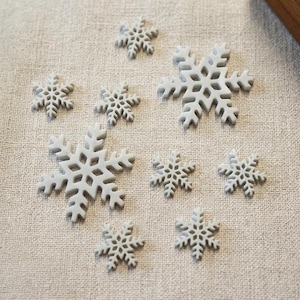 May include: A collection of eight light gray snowflake ornaments of varying sizes. Each snowflake features a detailed, symmetrical, laser-cut geometric pattern with six main branches and intricate, branching arms radiating from a central point.