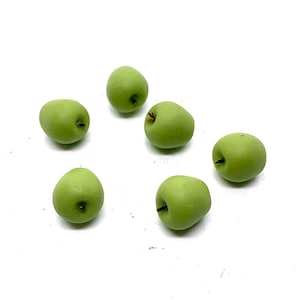 May include: Six small, green apples with dark brown stems are scattered on a white background. The apples have a matte finish and a slightly irregular shape, suggesting they are handmade or stylized.