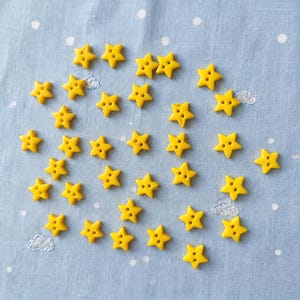 May include: A collection of small, bright yellow star-shaped buttons, each featuring two central holes for attachment. The buttons are scattered across a light blue fabric surface that is patterned with small, white, embroidered polka dots.