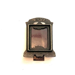 May include: A small, antique silver-toned picture frame with a rectangular opening and a decorative top. The frame has a dark, aged appearance and a small clasp at the bottom. The glass inside the frame is a muted purple color.