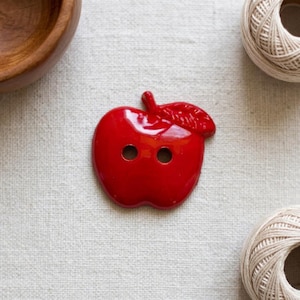 May include: A glossy, bright red apple-shaped button with a smooth finish and a textured leaf detail at the top. The button features two circular holes in the center for attachment, showcasing a vibrant, reflective surface and a distinct fruit silhouette.