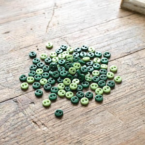 May include: A collection of small, round, two-hole buttons in various shades of green, ranging from light lime to deep forest green. These circular fasteners are scattered in a loose pile, each featuring a smooth surface and two central holes.