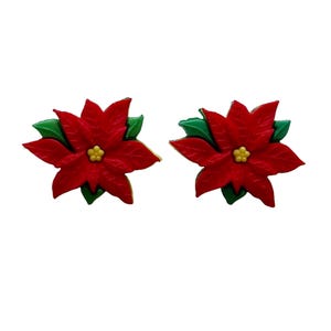May include: Two poinsettia flower earrings. Each earring features a vibrant red flower with green leaves and a yellow center. The earrings are set against a white background, highlighting the festive colors and design.