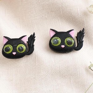 May include: Two black cat-shaped pins or charms featuring large, glossy green eyes, small pink noses, and pointed ears with pink inner details. Each cat figure has a curved, textured tail extending to the side, crafted with a smooth finish.