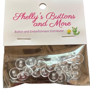 May include: A package of clear, shank buttons in a plastic bag. The buttons are round with a flat face and a loop on the back. The packaging has the text "Shelly's Buttons and More" in pink script.
