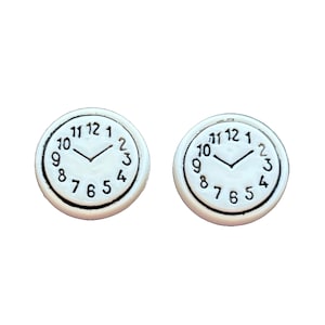 May include: Two white clock-shaped earrings. Each earring features a black-outlined clock face with the numbers 1 through 12. The hands of the clock are black and point to approximately 10:08. The earrings are set against a white background.