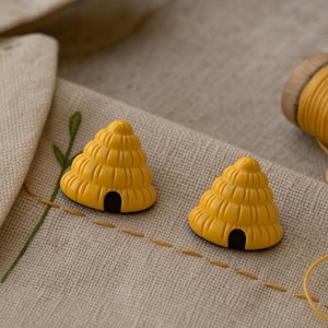 May include: Two yellow, cone-shaped beehive buttons with horizontal ridges and a small black arched entrance at the base. The buttons are placed on a piece of light beige fabric featuring simple green and brown embroidered stitching details.