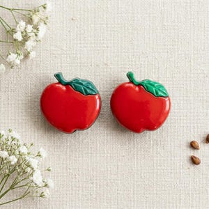 May include: A pair of enamel pins shaped like red apples with glossy green leaves. Each apple features a smooth, rounded red body and a single green leaf attached to the top stem, creating a vibrant and polished appearance for these accessories.