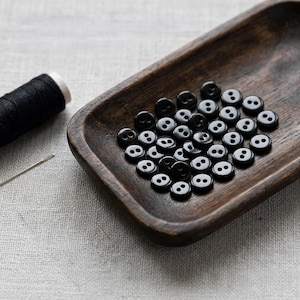 May include: A collection of small, round, black two-hole buttons resting inside a dark, elongated wooden tray. Beside the tray lies a spool of black thread and a single silver sewing needle, all arranged on a light-colored fabric surface.