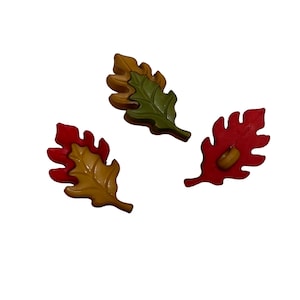 May include: Three leaf-shaped buttons in autumn colors. One button is green with a brown border, one is yellow with a red border, and one is solid red. Each button features a distinct serrated edge design and a central attachment point on the back.