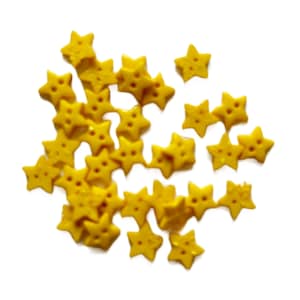 May include: A collection of small, yellow star-shaped sprinkles with three small holes in each star. The sprinkles are scattered on a white background, creating a playful and festive appearance. These are likely for decorating baked goods.
