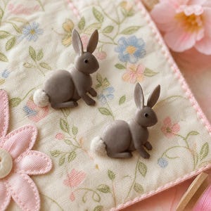 May include: Two small, grey rabbit-shaped buttons with white fuzzy tails, positioned on a cream-colored fabric square featuring a delicate floral pattern in pink, blue, and green, with a pink stitched border and a large pink fabric flower.