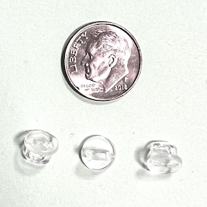 May include: Three small, clear, circular plastic buttons with a smooth, translucent finish. Each button features a central shank for attachment. A standard United States dime is positioned above the buttons to provide a clear scale reference.