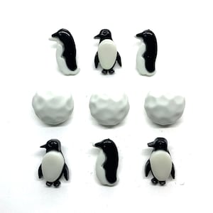 May include: Nine small decorative items: six penguins and three white spheres. The penguins are black and white, with a glossy finish. The spheres are white and textured, resembling snowballs. All items are arranged on a white surface.