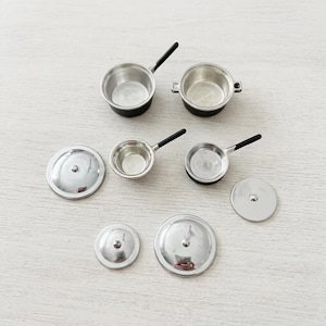 May include: A set of miniature kitchen cookware including two small pots with black handles, two small pans with black handles, and four matching silver-colored circular lids. The items are metallic silver with black accents on the handles.