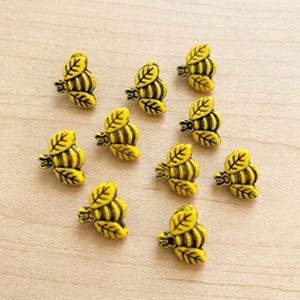 May include: A collection of nine small, yellow bee-shaped buttons. Each button features a detailed, embossed design with black accents highlighting the wings and body segments. The wings are shaped like leaves, and the bodies have horizontal stripes.
