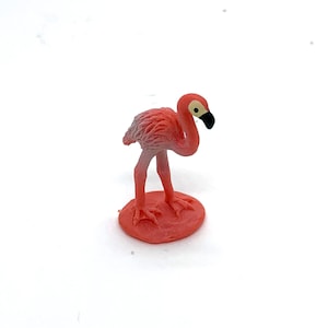 May include: A small, pink flamingo figurine with a black beak and yellow eye. The flamingo stands on a round, coral-colored base. The figurine is made of plastic and is set against a white background.