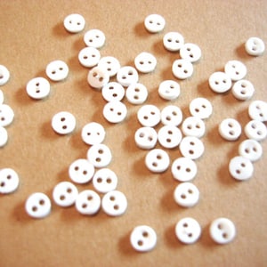 May include: A collection of small, white, two-hole buttons scattered on a brown surface. The buttons are round and uniform in size, with a smooth, glossy finish. Ideal for sewing projects, these buttons offer a classic and versatile design.