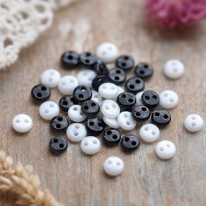 May include: A collection of small, round, two-hole buttons in solid black and white colors. These circular fasteners are scattered in a cluster, featuring a smooth finish and a central indentation for the two holes used for sewing attachment.