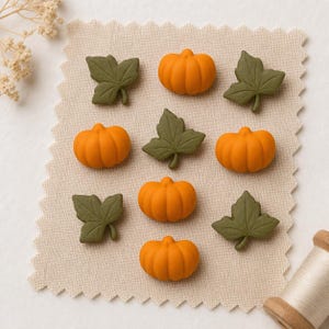May include: A collection of small, orange pumpkin-shaped embellishments and olive green leaf-shaped embellishments arranged in a grid pattern on a piece of light beige fabric with pinked edges, featuring four pumpkins and four leaves.