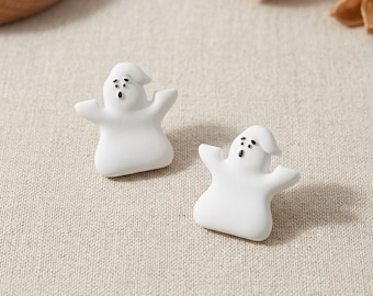 May include: Two small, matte white ghost-shaped figurines. Each ghost features a simple, rounded body with outstretched arms and a hand-painted face consisting of two small black dots for eyes and a single black dot for an open, surprised mouth.