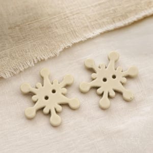 May include: Two beige buttons shaped like six-pointed snowflakes with rounded tips. Each button features a smooth, matte finish and two central holes for attachment, displaying a consistent light tan color throughout the entire structure.