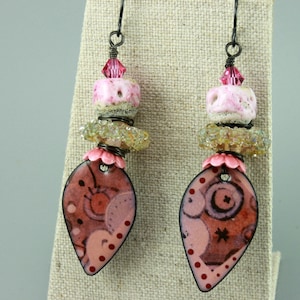 May include: A pair of handcrafted earrings with a unique design. Each earring features a teardrop-shaped pendant with a pink and brown pattern, accented with red dots. The earrings are adorned with pink and clear beads and a pink crystal.