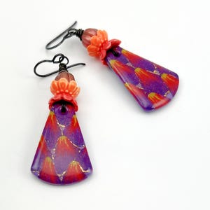 May include: A pair of handmade earrings with a floral theme. The earrings feature a purple, red, and gold pattern with a flower accent. The earrings are approximately 2 inches long.