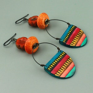 May include: A pair of handcrafted earrings featuring orange and red beads with a dark metal hook. The earrings have a teardrop-shaped pendant with a teal background and colorful stripes in red, yellow, and orange.